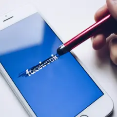 Dłoń z rysikiem próbuje sprawdzić, jak sprawdzić ile zostało blokady na FB, dotykając ekranu telefonu z logo Facebooka.
