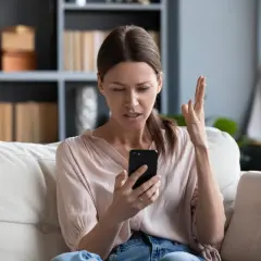 Zirytowana kobieta na kanapie widzi na telefonie komunikat: brak dostępnej sieci komórkowej umożliwiającej wykonywanie połączeń głosowych.