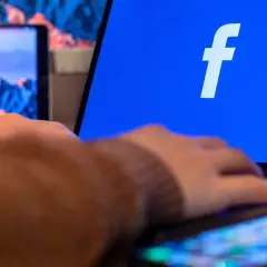 Dłoń na klawiaturze laptopa, na ekranie logo Facebooka. Dowiedz się, jak utworzyć stronę na Facebooku.