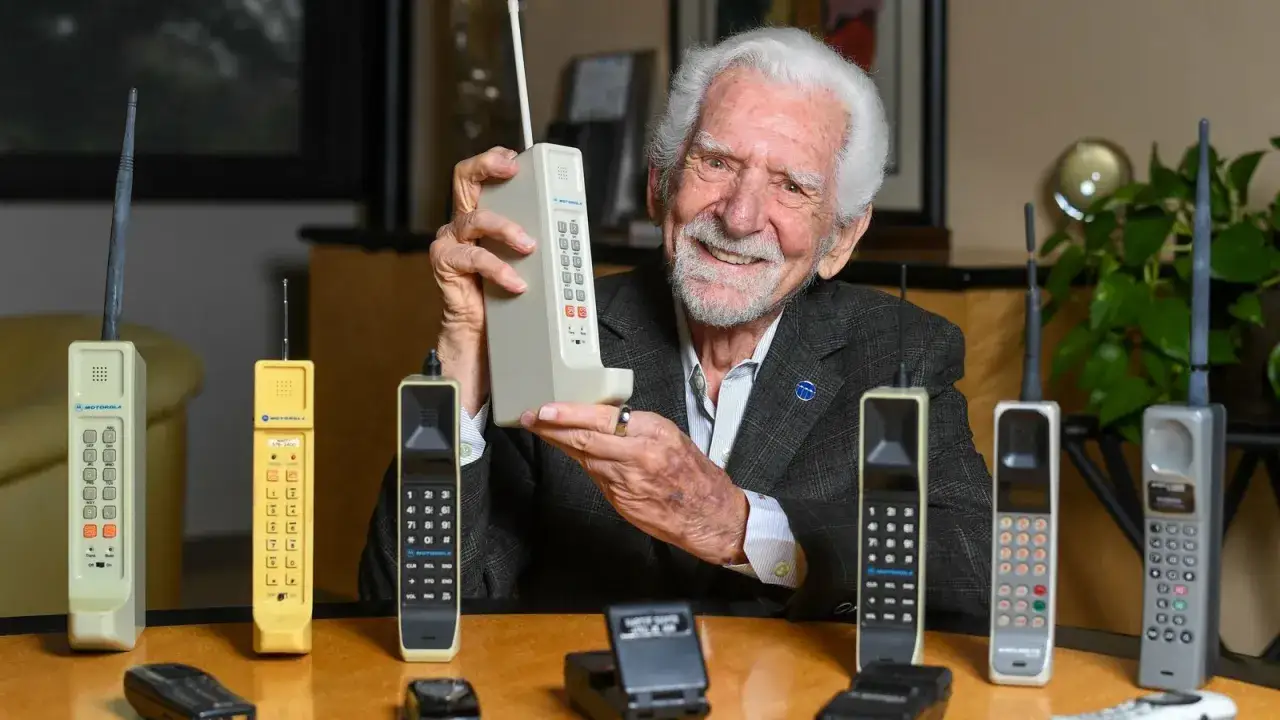 Uśmiechnięty starszy pan z siwą brodą trzyma pierwszy telefon komórkowy, pokazując ewolucję technologii od czasów, kiedy powstał pierwszy telefon komórkowy.
