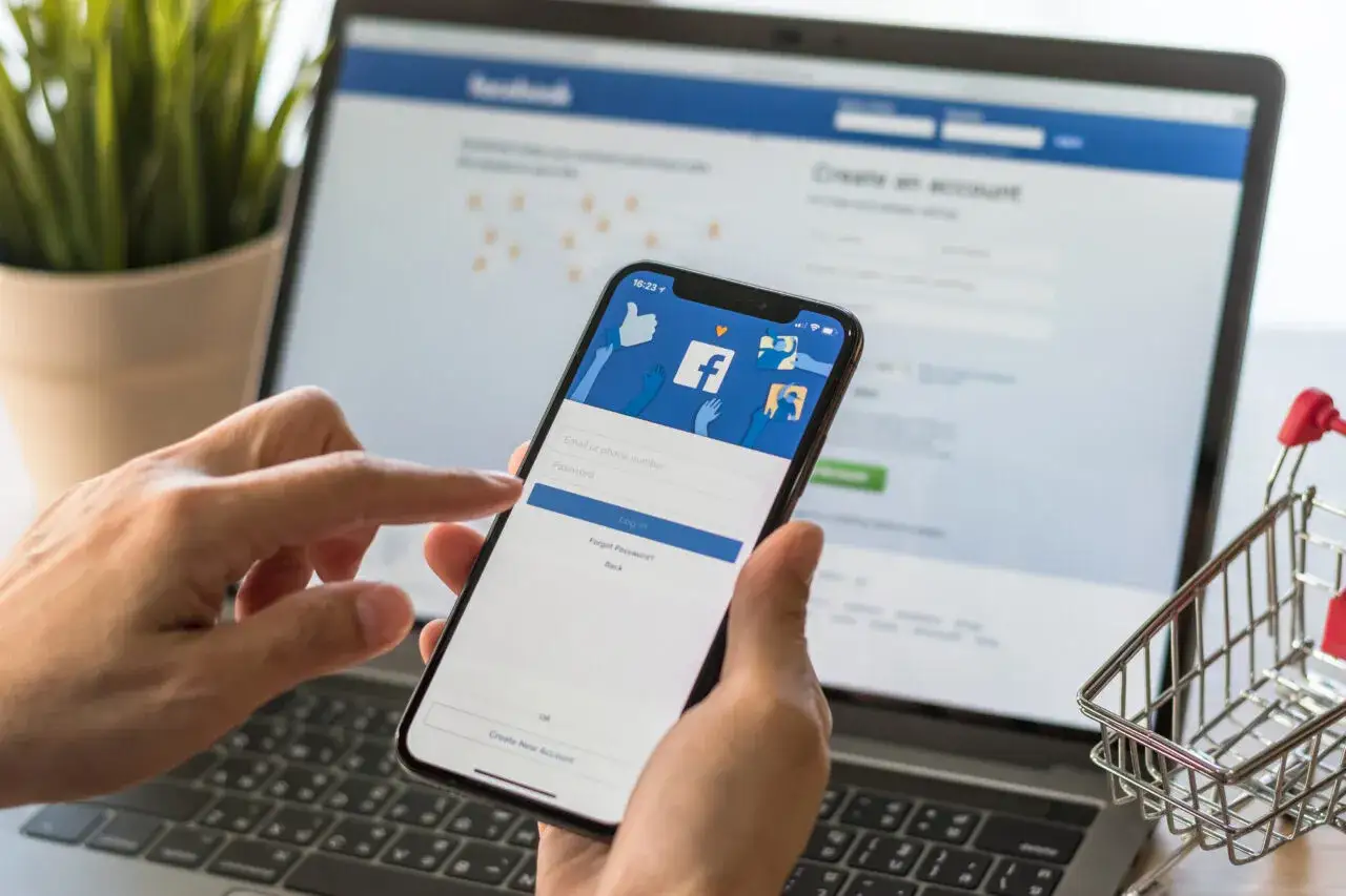 Ręce trzymające smartfon z ekranem logowania Facebooka. Obok laptop z otwartą stroną FB. Jak sprawdzić kto jest administratorem strony na FB?