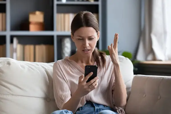 Zirytowana kobieta na kanapie widzi na telefonie komunikat: brak dostępnej sieci komórkowej umożliwiającej wykonywanie połączeń głosowych.