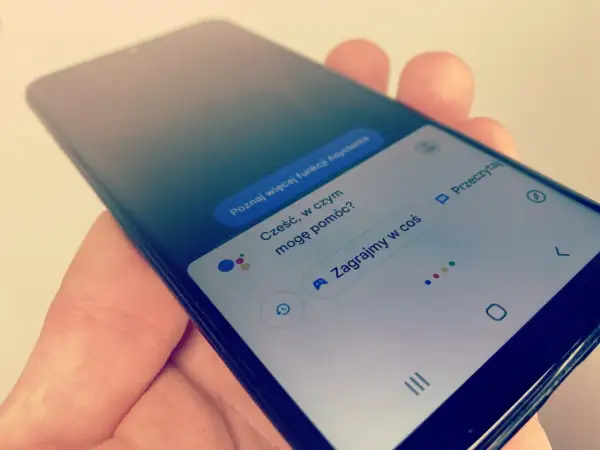 Smartfon z Asystentem Google, który czeka na **wyszukiwanie głosowe**. Widoczne opcje: "Poznaj więcej funkcji Asystenta", "Cześć, w czym mogę pomóc?", "Zagrajmy w coś".